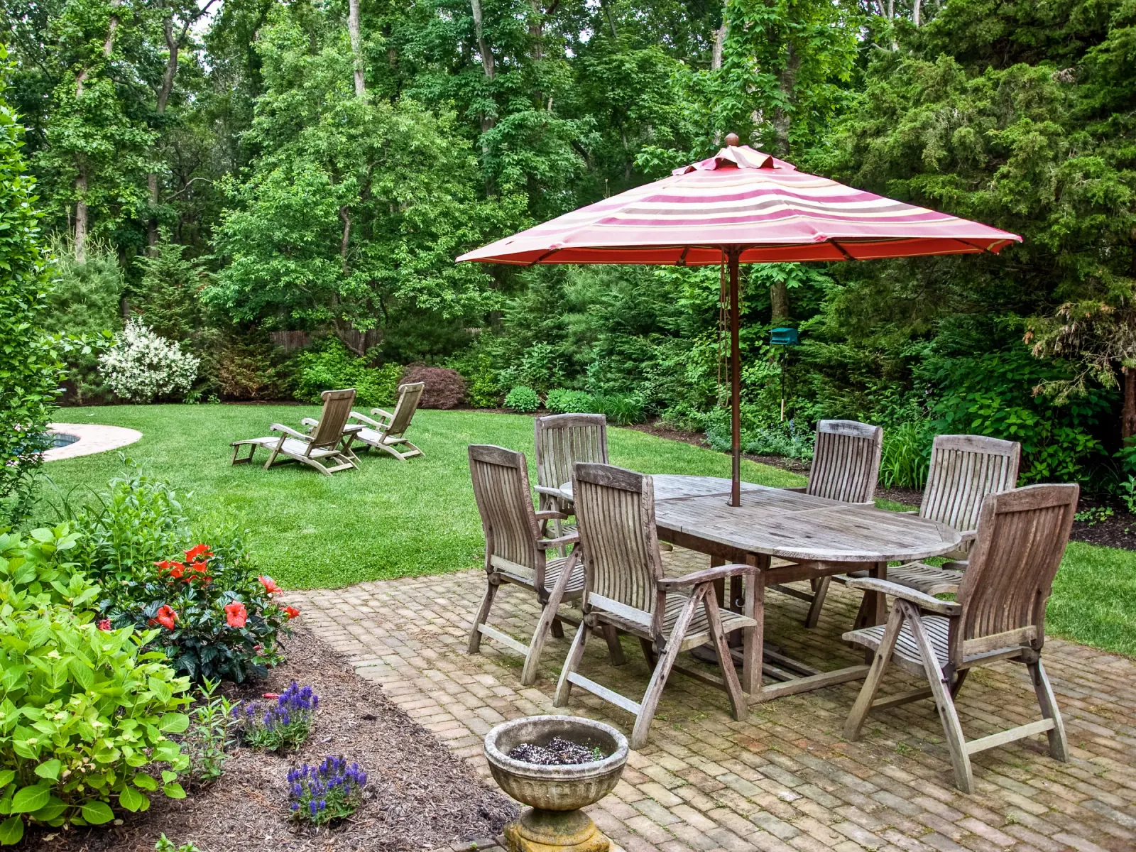 A clean back yard with a brick patio, table and umbrella, and a wide lawn bordered by trees